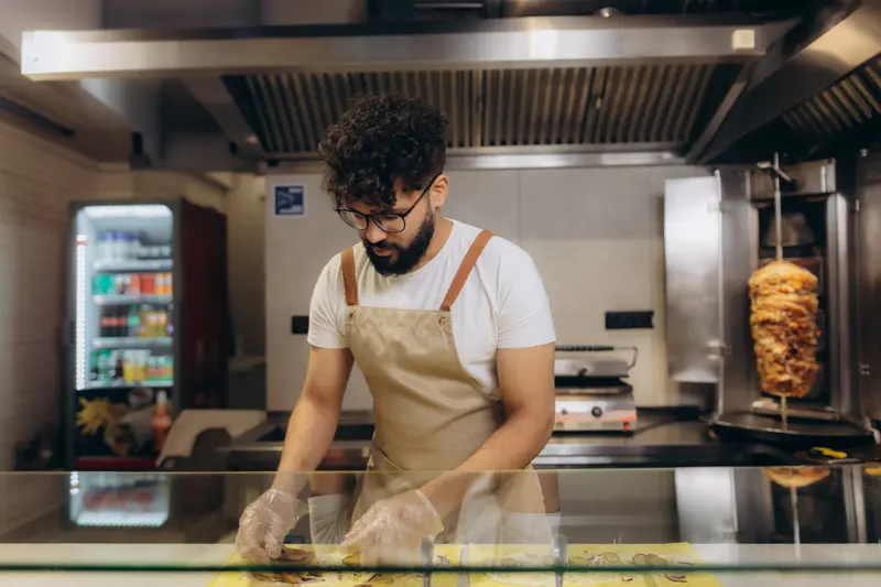 Grillkock i förkläde skär kött från kebabspettet i ett ljust kök