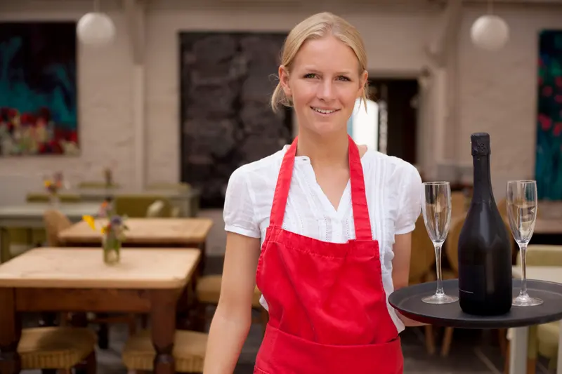 Servitris i röd förkläde bär en bricka med glas genom restaurangen