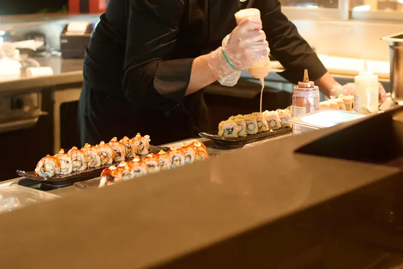 Sushikock lägger sås på en rad färdiga rullar vid disken